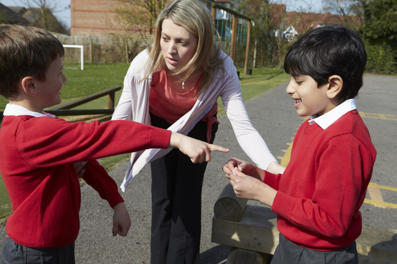 Teacher talking to two primary school pupils. One pupil is pointing at the other. 